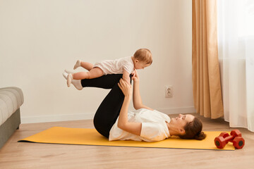 Attractive young mother doing sport exercises together with her infant baby, woman lying on floor on yoga mat and playing with her cute toddler kid while having workout at home.