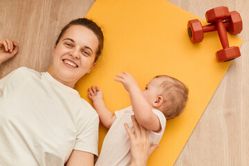 Obraz na płótnie Canvas Top view of young mother with her baby daughter lying on the fitness mat during the exercise with dumbbells on the floor, resting, looking smiling at camera.