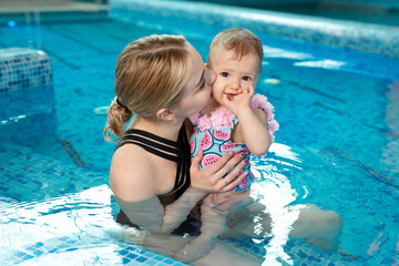 A young mother holds her little daughter in the pool, hugs her and kisses her.