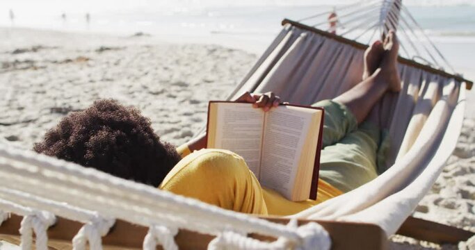 African american woman reading and lying in hammock on sunny beach