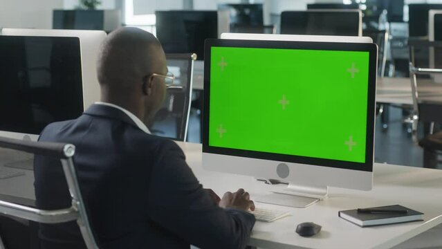 African-American Business Man Typing on Desktop Computer with Green Mock-up Screen while Sitting at His Desk. In the Background Stylish Modern Open Space Creative Business Office