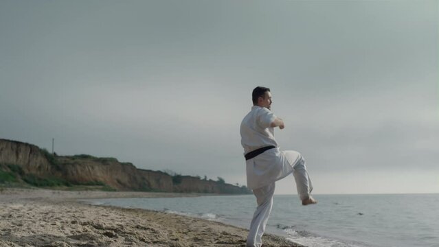 Karate athlete practicing kicks on sunny beach. Sporty man training martial arts