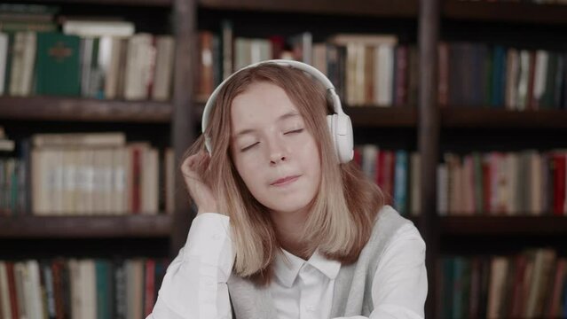teenager with short hair wearing pink sweater listening to favorite music with headphones while sitting in library