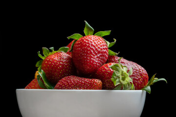 Ripe red strawberries with green tails in a white plate on a black background. Sweet dessert of fresh berries on the table.