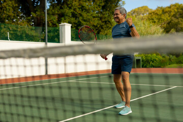 Obraz premium Cheerful biracial senior man holding racket gesturing while playing tennis in court seen through net