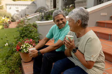 Smiling biracial senior man talking with senior wife holding coffee cup while sitting on steps - Powered by Adobe
