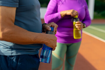 Midsection of biracial senior man and woman opening water bottles while standing in tennis court - Powered by Adobe
