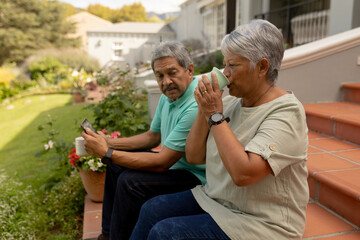 Biracial senior woman drinking coffee and senior man using digital tablet while sitting on steps