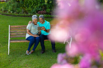 Fototapeta premium Cheerful biracial senior man with wife using digital tablet while sitting on bench in park