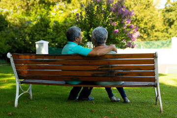 Biracial senior husband with hand on senior wife's shoulder sitting on bench against trees in park