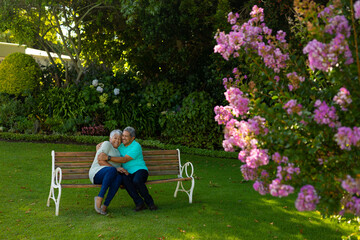 Romantic biracial senior man embracing wife while sitting on bench against trees and plants in park - Powered by Adobe