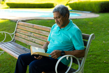 Fototapeta premium Focused biracial senior man wearing blue t-shirt reading book while sitting on bench in park
