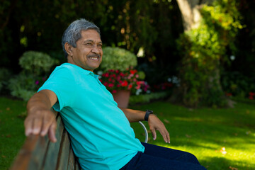 Smiling biracial senior man looking away while sitting on bench against plants in park - Powered by Adobe