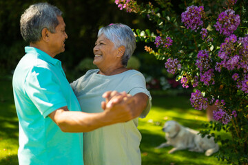 Side view of smiling biracial senior couple looking at each other while dancing by flowers in park - Powered by Adobe