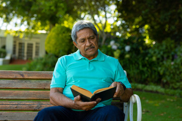 Obraz premium Serious biracial senior man reading book with focus while sitting on bench against plants in park
