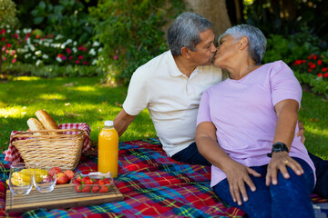 Biracial senior couple kissing while sitting with bread, fruits and juice on blanket in park