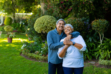 Smiling biracial senior couple embracing and looking up while standing against plants in park - Powered by Adobe