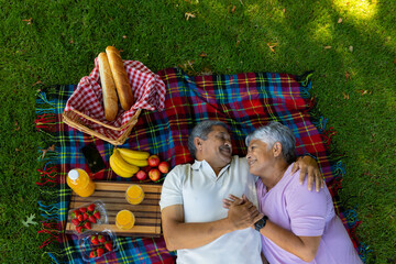 High angle view of smiling biracial senior couple with food and drink lying on blanket in park