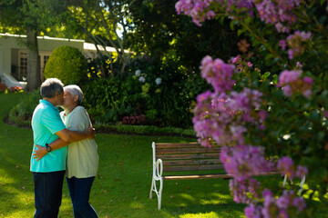 Side view of biracial senior couple embracing and kissing while standing against plants in park - Powered by Adobe