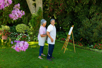 Biracial senior woman tying husband's apron standing by canvas and and easel against plants in yard - Powered by Adobe