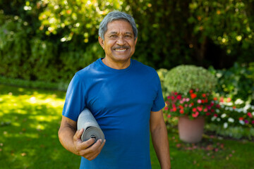 Portrait of smiling biracial senior man in sports clothing holding yoga mat against plants in yard