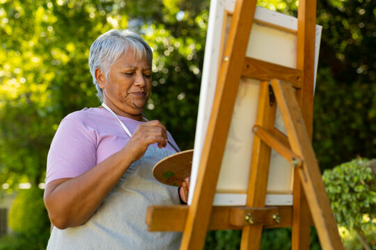 Low angle view of biracial senior woman with short hair painting with watercolors on canvas in yard