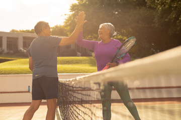 Cheerful biracial senior couple giving high-five while playing tennis at court during sunset - obrazy, fototapety, plakaty