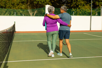 Rear view of biracial senior husband and wife with arms around walking at tennis court on sunny day - Powered by Adobe