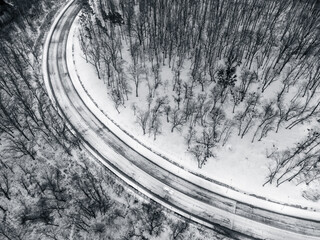 Fly above winter snowy road curve. Black and white highway surrounded by forest covered in snow. Aerial look down view