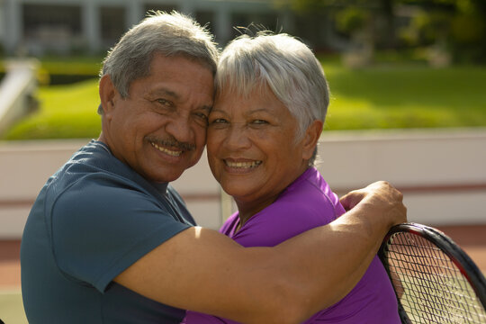 Close-up portrait of smiling biracial senior husband embracing senior wife at tennis court