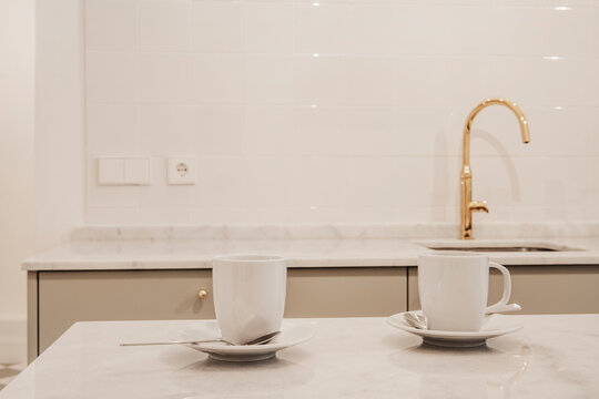 Pair Of White Coffee Mugs On White Marble Countertop In A Kitchen With Gold Taps