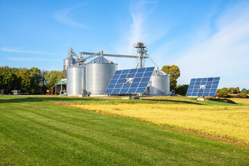 Solar panels and a grain elevator with big steel storage bins in a modern farm on a sunny autumn...