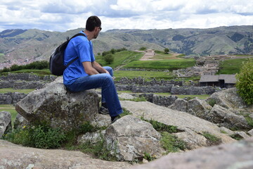 A man sits on a rock and looks at ruins of saqsaywaman, Cuzco. Peru