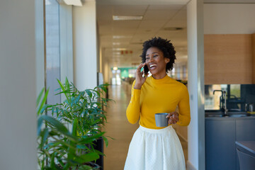 Cheerful young african american businesswoman talking on smartphone holding coffee mug at workplace