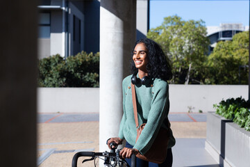 Fototapeta na wymiar Smiling young biracial businesswoman walking with bicycle on sunny day