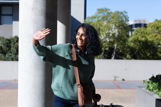 Cheerful young biracial businesswoman standing with bicycle while waving hand on sunny day