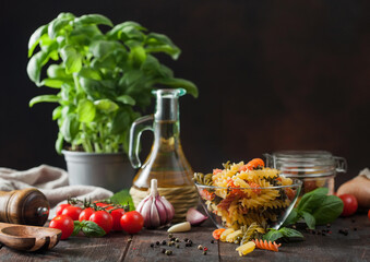 Raw tricolor fusilli pasta in glass bowl with oil and garlic, basil plant and tomatoes on wooden background.