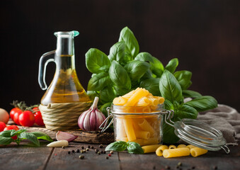 Raw fresh penne pasta in glass jar with oil and garlic, basil plant on wooden background.