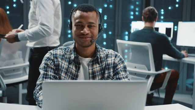 Portrait of young smiling African man wearing checkered shirt sitting at desktop with laptop. Footage of friendly male call center operator in headset with microphone looking directly at camera