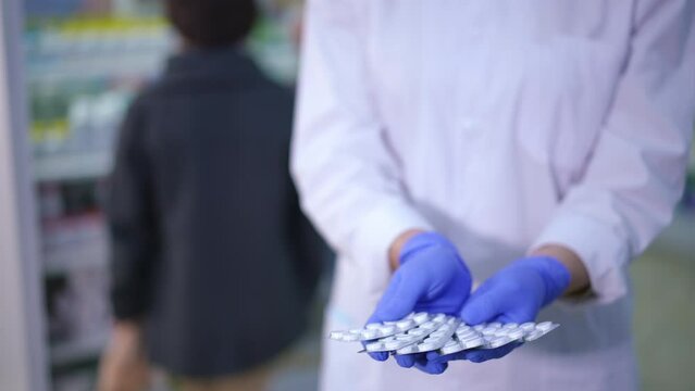 Front view unrecognizable druggist stretching white pills in blisters to camera standing in drugstore with blurred customers at background. Young Caucasian woman advertising drugs in pharmacy indoors