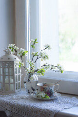 Fototapeta na wymiar A cup of tea, a cherry branch in a glass vase stand on the window. Spring still life in vintage style.