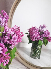 A bouquet of lilacs in a vase reflected in a round mirror on a gray background.
