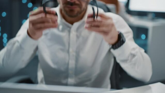 Footage of serious confident young bearded man in white shirt sitting at desktop, wearing headphones. Male call center operator talking to client, discussing problem, consulting. Blurred background