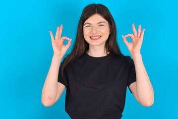 Glad young caucasian woman wearing black T-shirt over blue background shows ok sign with both hands as expresses approval, has cheerful expression, being optimistic.