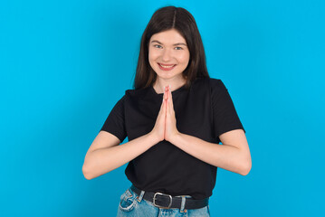 young caucasian woman wearing black T-shirt over blue background keeps palms together, has pleased...