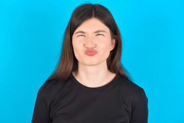 young caucasian woman wearing black T-shirt over blue background crosses eyes, puts lips, makes...