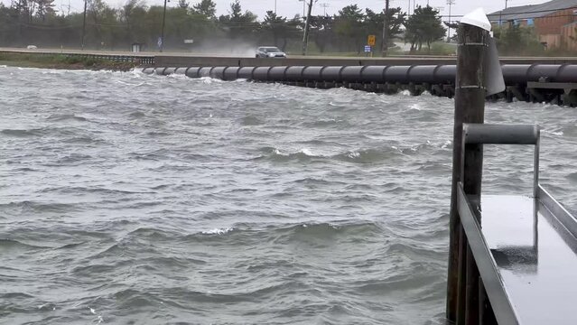 Rough bay water crashes against bulkhead, road and large oil pipeline as cars rush by during a storm