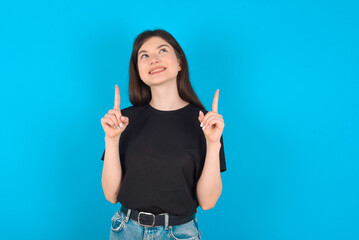 Successful friendly looking young caucasian woman wearing black T-shirt over blue background exclaiming excitedly, pointing both index fingers up, indicating something.