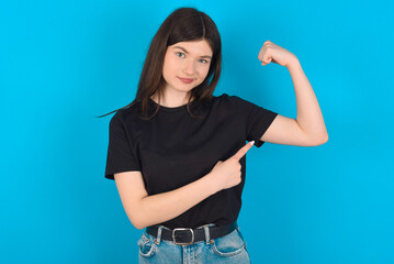 Smiling young caucasian woman wearing black T-shirt over blue background raises hand to show muscles, feels confident in victory, strong and independent.