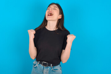 Fototapeta na wymiar young caucasian woman wearing black T-shirt over blue background looks with excitement up, keeps hands raised, notices something unexpected.